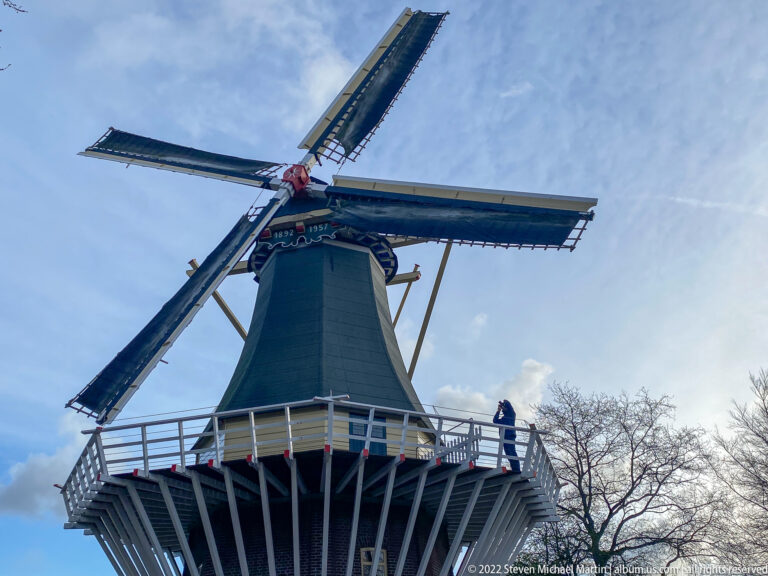 Upward centered view of the Keukenhof windmill with four sails, raised gallery, and a visitor against the sky.