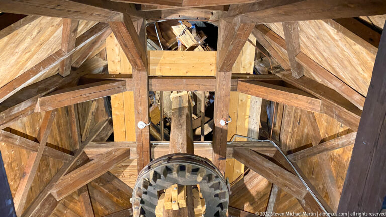Interior of a wooden windmill showing heavy timber framing, a large toothed gear, shafts, braces, and reinforced structural members.