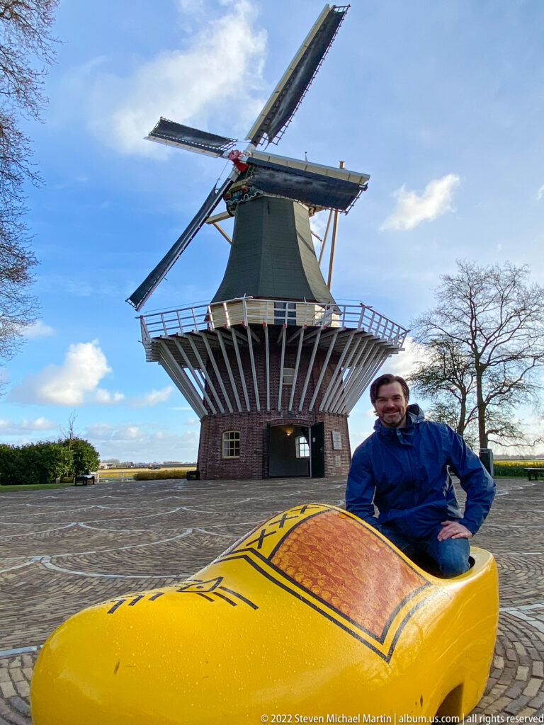 Keukenhof windmill behind a large yellow decorative Dutch clog, with a visitor standing on the cobbled forecourt.