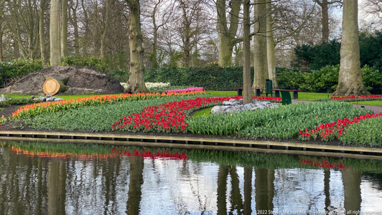 Red and orange tulip beds line a narrow canal, with their colors reflected in the water and a cut tree trunk behind.