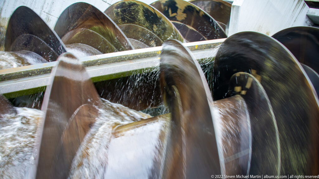 Close view of large helical screw pumps churning water within a metal-lined channel.