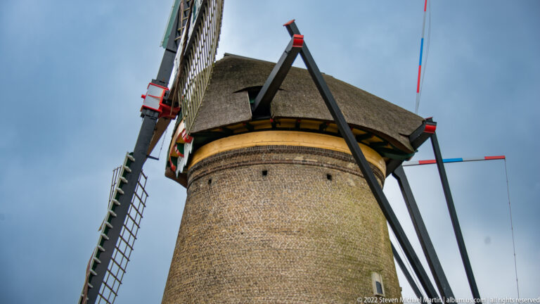 Low-angle close view of a brick windmill tower, thatched cap, sails, and painted structural poles.