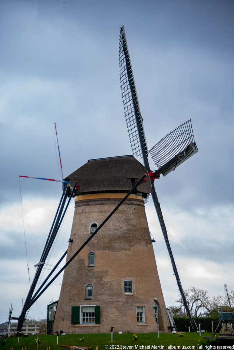 Large brick windmill seen broadside with thatched cap, sails, windows, and long support braces.