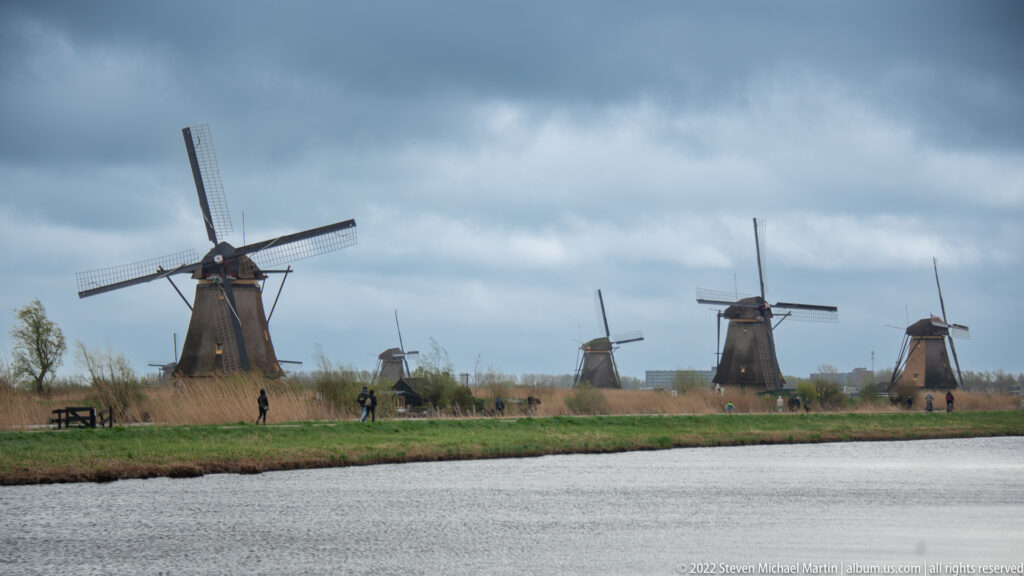 Several windmills beyond a canal, with small people walking along the embankment path.