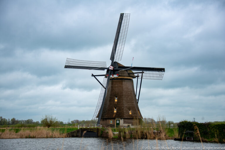 Dark timber-clad windmill beside water with a small arched crossing nearby.