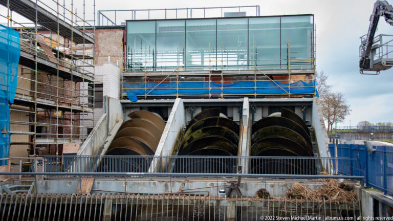 Three large screw pumps beneath a glass-fronted building with scaffolding and construction materials.
