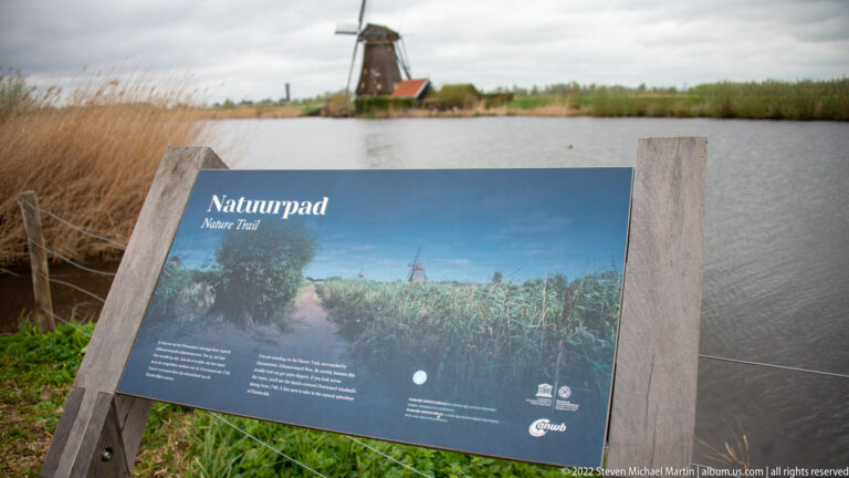 Nature Trail interpretive panel beside water, with reeds and a windmill in the background.