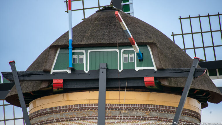 Close view of a brick windmill’s green-painted upper structure, thatched cap, and crossing sail supports.