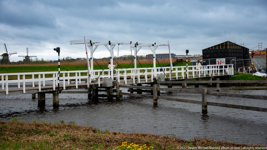 White timber footbridge spanning a narrow waterway, with windmills visible in the distance.