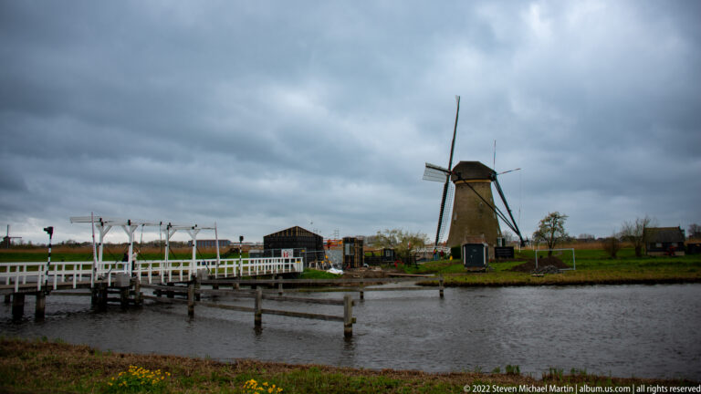 White footbridge crossing a waterway with a windmill and visitor buildings on the far bank.