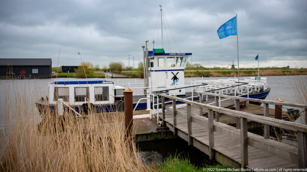 White passenger boat moored beside a wooden landing and reeds, with a blue flag above it.