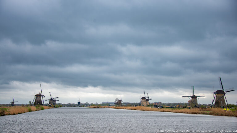 Broad canal stretching between rows of windmills beneath dark storm clouds.
