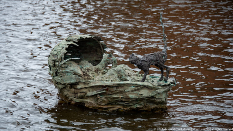 Weathered bronze cradle-like sculpture in the water with a small bird figure perched on one side.
