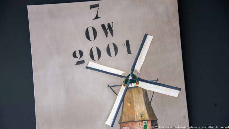 Small model windmill mounted on a wall beneath black stenciled text reading OW 2001.