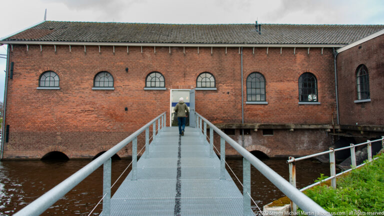 Person walking across a metal footbridge toward a long red-brick industrial building over the water.