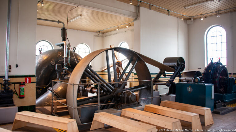 Large spoked flywheel and historic pumping machinery inside a bright industrial room with arched windows.