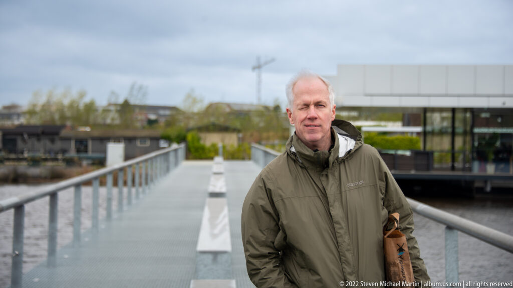 Adult man in a dark jacket standing on a long pedestrian bridge over water with visitor buildings behind him.