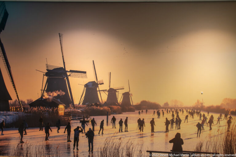 Historic winter scene of many people skating on frozen water beneath several windmills at sunset.