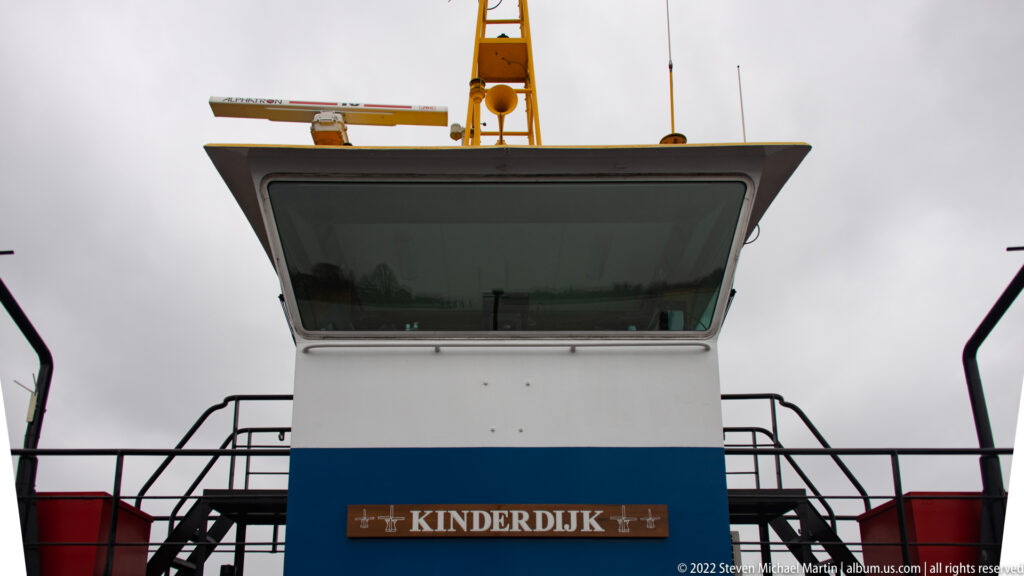 Front of a white-and-blue vessel wheelhouse with a wooden KINDERDIJK nameboard below the windows.
