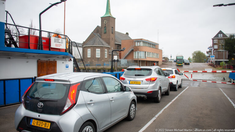 Cars queued beside a vessel and red-white barriers, with a brick church and green spire in the background.