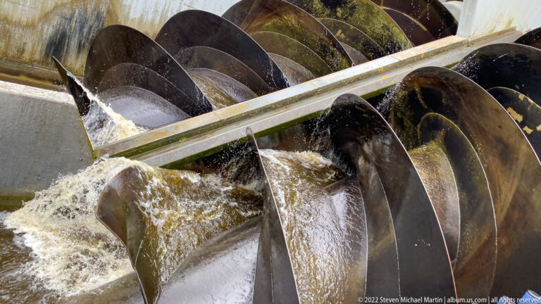Close view of large black helical screw pumps lifting turbulent water beside a metal divider.