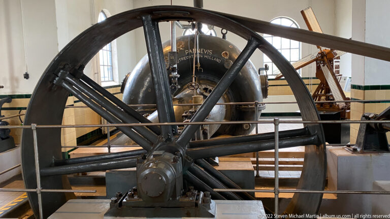 Large black spoked flywheel in front of a rounded Pannevis vessel and associated pumping machinery.