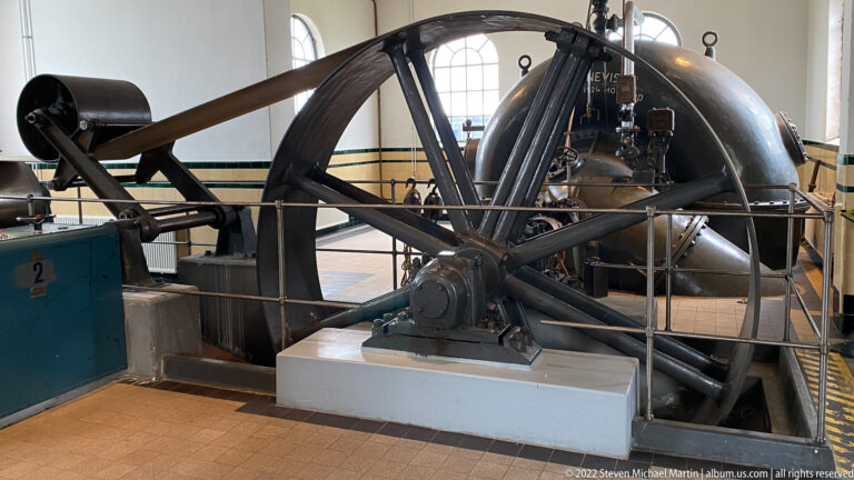Wide interior view of a large spoked flywheel, Pannevis vessel, shafts, railings, and adjacent pumping equipment.