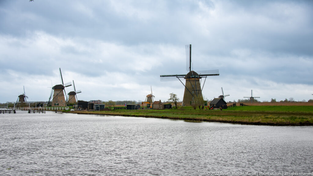 Several windmills spread across flat fields beyond a broad canal under a cloudy sky.
