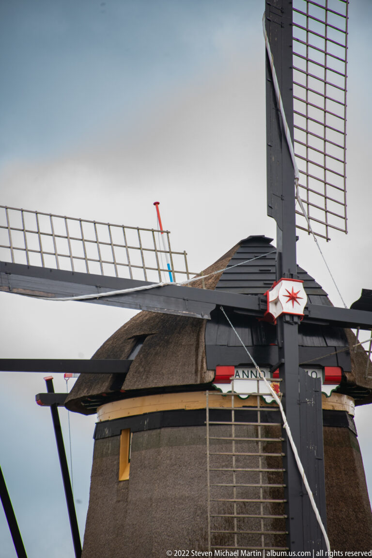 Close view of a windmill’s latticed sails, dark cap, and upper brick tower with painted trim.