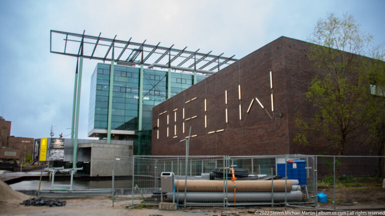 Brick museum building with narrow vertical openings, glass tower, steel canopy, fencing, and exposed pipes.
