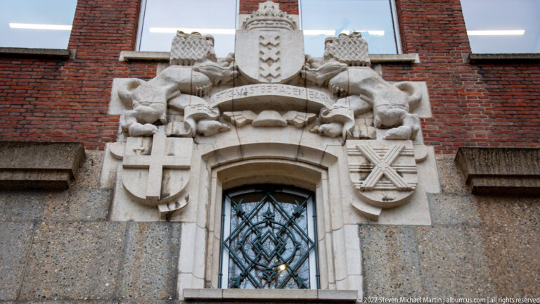 Pale stone heraldic crest with shields and carved figures above an arched window in a red brick façade.