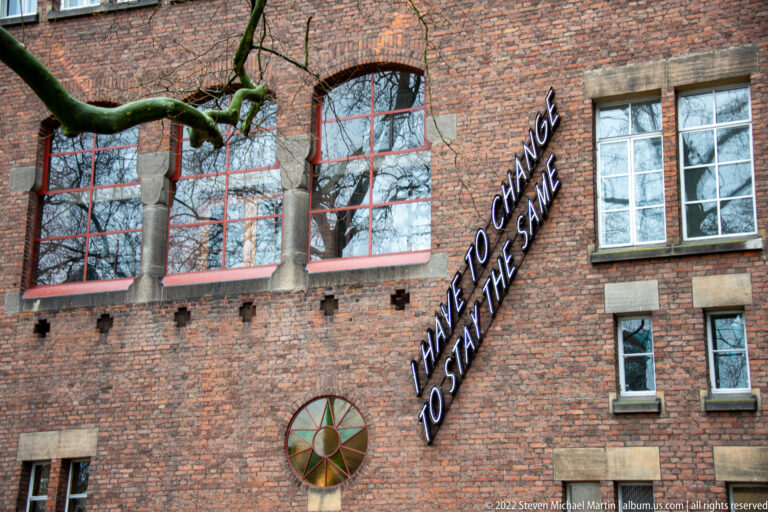 Brick façade with multi-pane windows, a tree, circular wall ornament, and illuminated text reading “I have to change to stay the same.”