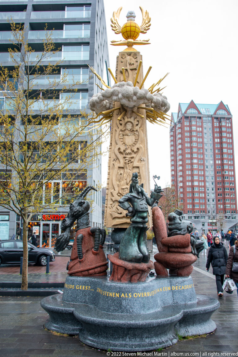 Stone and bronze Rotterdam monument with sculpted figures, reliefs, gilded emblem, and the motto “Sterker door strijd.”