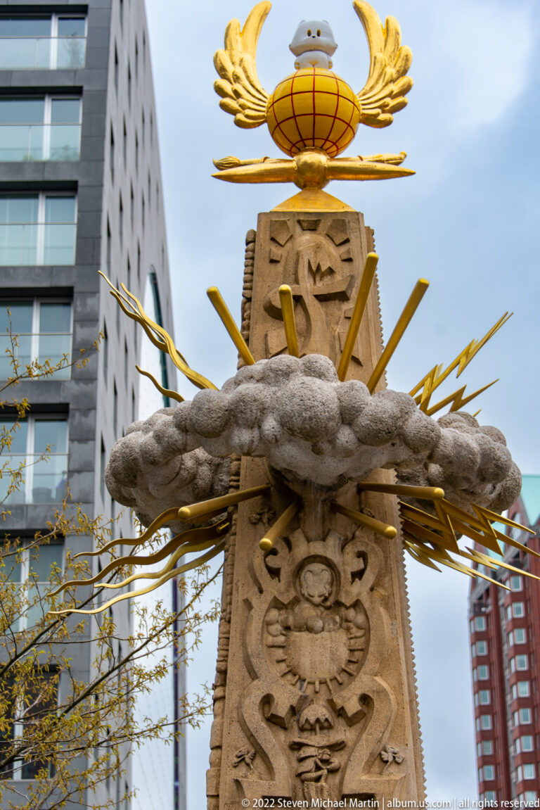Close view of gilded winged globe, radiating gold rods, gray sculptural band, and carved stone monument shaft.