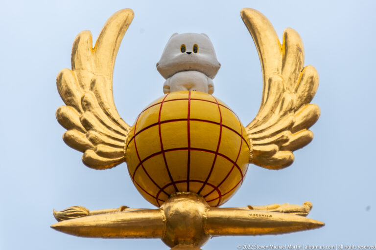 Gilded winged globe with small pale figure above and a horizontal gold element against blue-gray sky.
