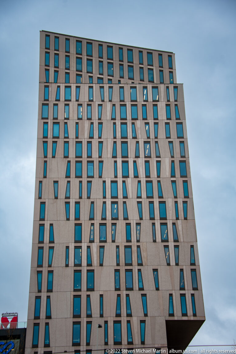 Tall stone-toned high-rise with irregular dark windows, narrow vertical recesses, and a subtly tapering façade.