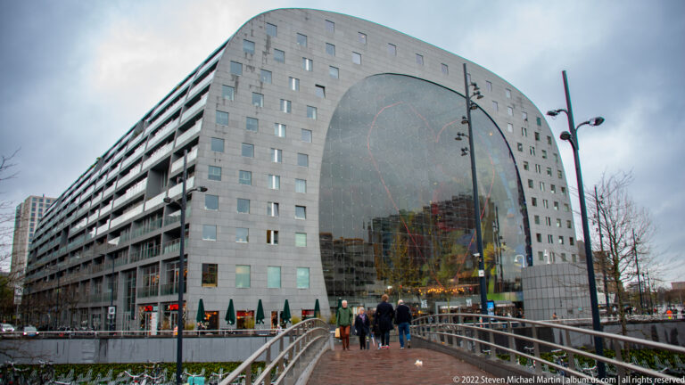 Markthal Rotterdam's horseshoe-shaped stone façade and huge glazed end wall seen from a pedestrian bridge with people walking toward it.