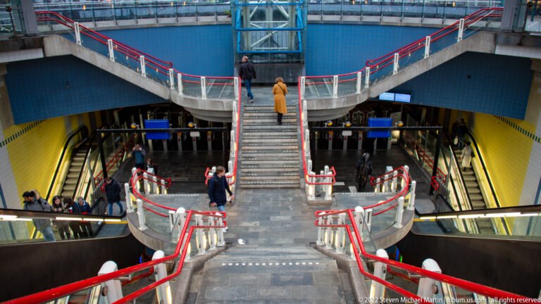 Blaak metro station with split stairs, escalators, red handrails, blue walls, yellow lighting, and commuters.
