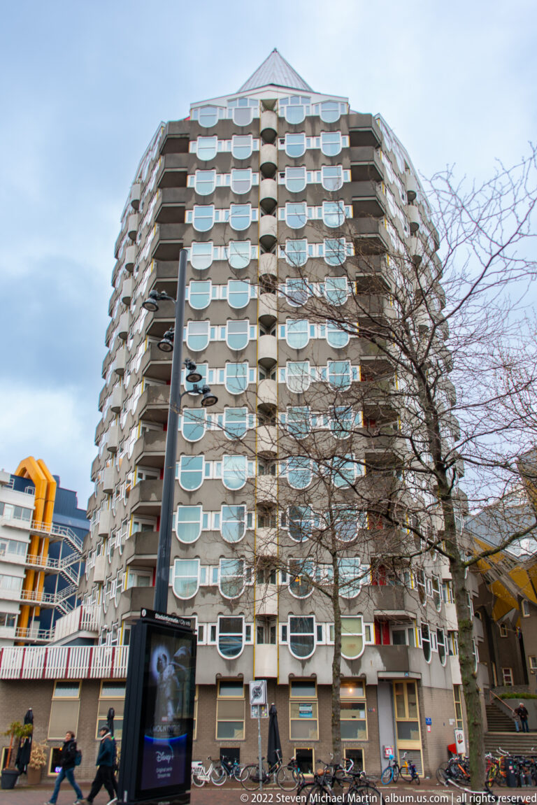 Rounded apartment tower with wraparound balconies and repeating white-framed windows above bicycles and street activity.