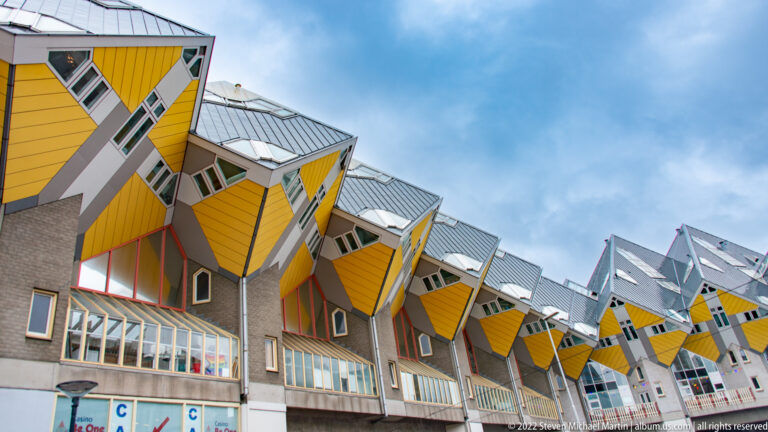 Long row of yellow Cube Houses receding above pedestrian-level windows and passages beneath a blue sky.