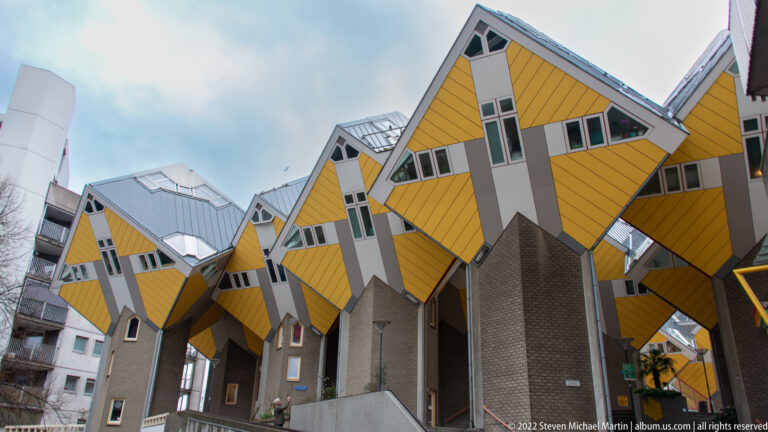 Clustered rear and side views of yellow Cube Houses on tall brick bases, with narrow passages and deep overhangs.