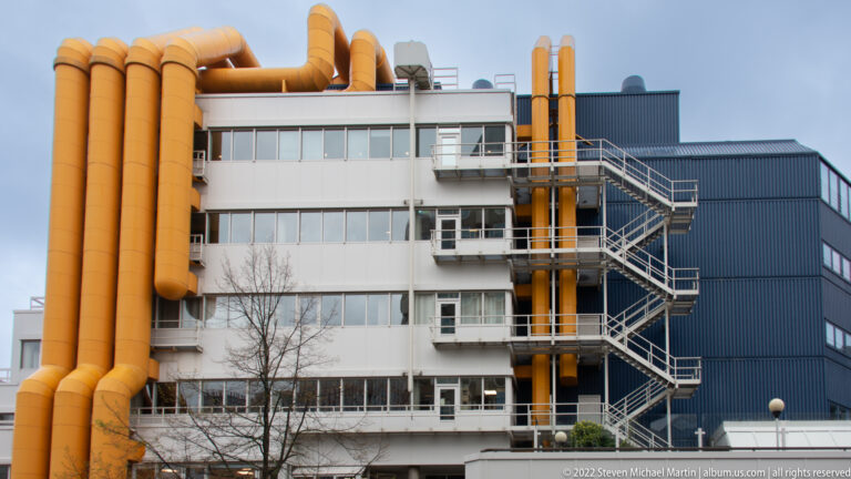 Central library façade with large yellow exterior ducts, white horizontal bands, metal fire stairs, and a dark blue upper volume.