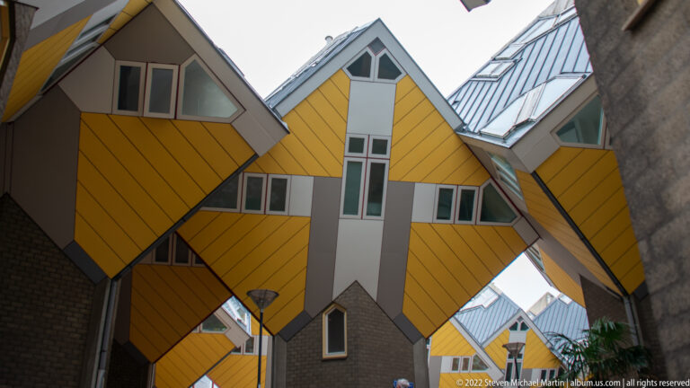 Upward view beneath yellow tilted Cube Houses, with angular windows, gray roofs, brick bases, and a narrow passage below.