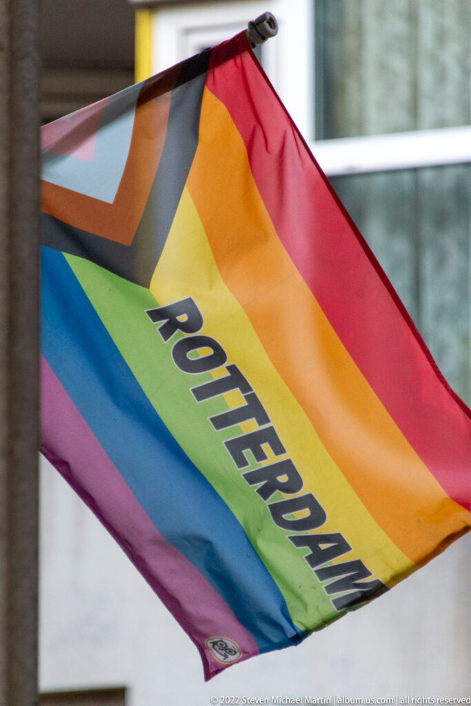 Rainbow Pride flag blowing in the wind with the word ROTTERDAM printed diagonally across the colored bands.