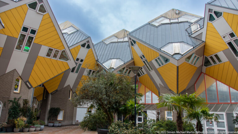 Courtyard with palms and potted plants beneath yellow Cube Houses, surrounded by tilted walls, windows, and gray roof planes.
