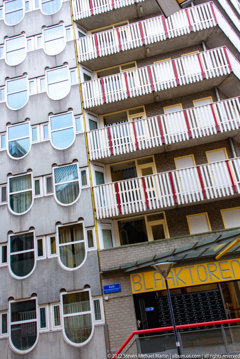 Angled view of adjoining apartment façades with rounded projecting windows and long balconies shaded by red-and-white striped awnings.