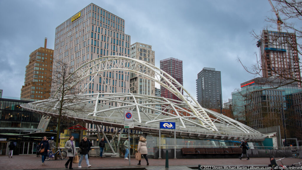 White ribbed station canopy arching above pedestrians and bicycles, with high-rise buildings and construction cranes behind.