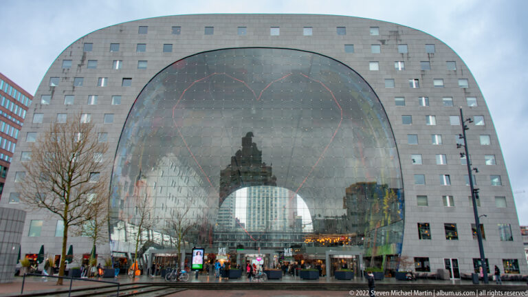 Large horseshoe-shaped Markthal façade with a glass arched entrance, small apartment windows, pedestrians, and reflected city buildings.