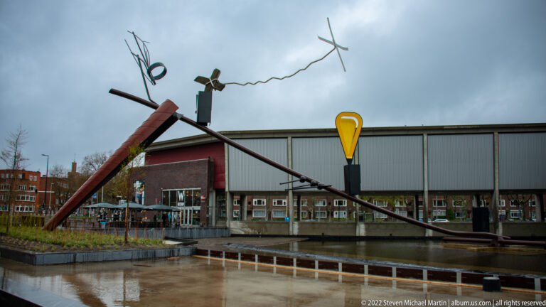 Diagonal red and black abstract sculpture with yellow center on a wet plaza before a low cultural building.