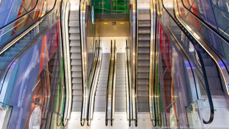Two escalators descending side by side through a modern interior with glass rails, polished metal, and colorful reflections.